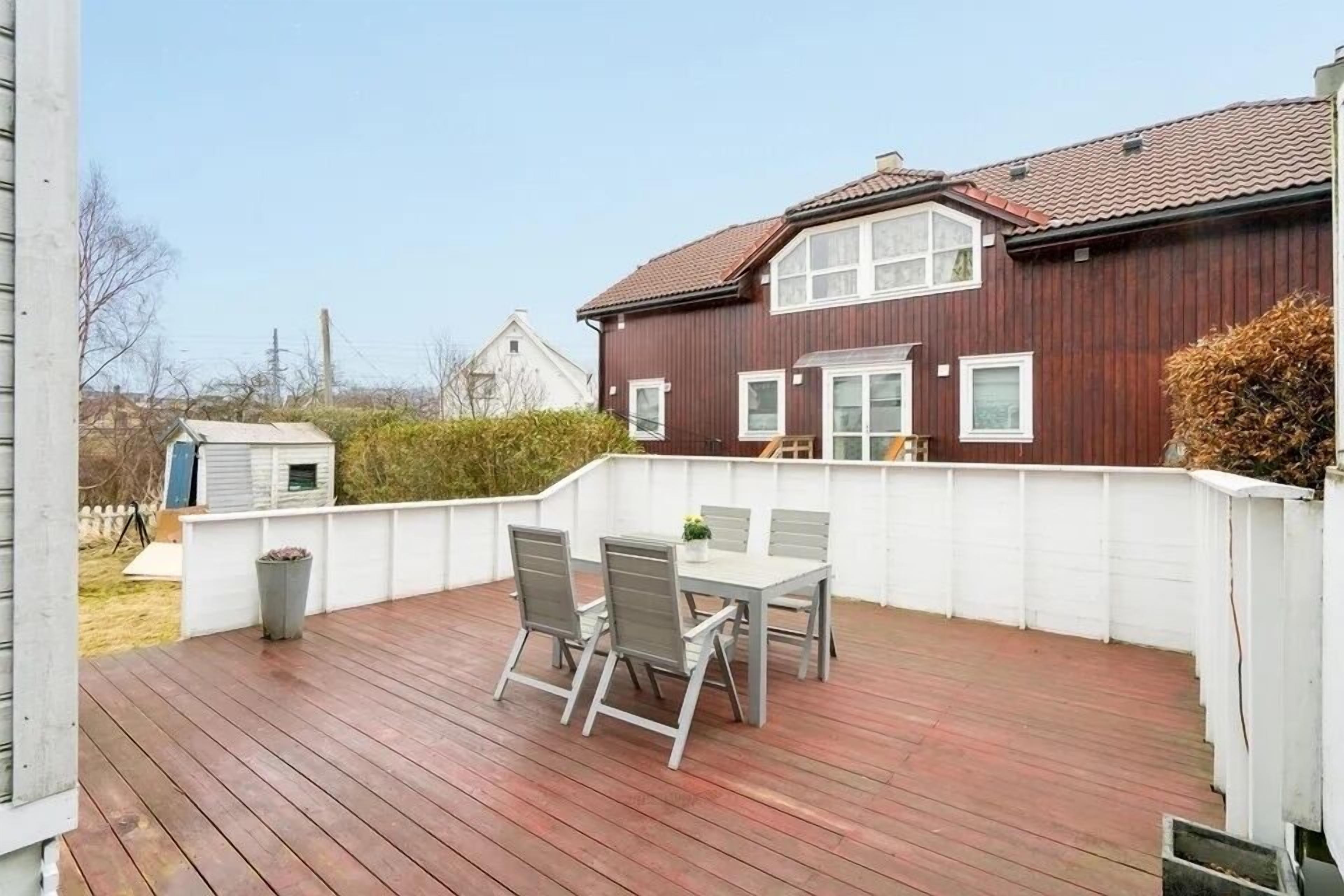 A wooden deck with a table and chairs, a white fence, and a house with a red roof in the background.