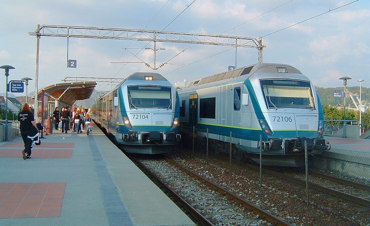 Dos trenes en el andén de una estación, con líneas eléctricas elevadas y una zona de espera cubierta.