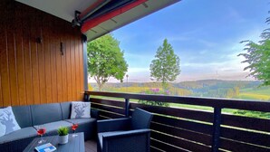 A balcony with wicker furniture, a glass table, and a view of a forested landscape.