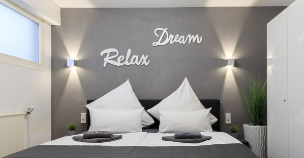 Et moderne soveværelse med en grå væg med struktur, to hvide puder, en sort sengegavl og et skilt med teksten 'Dream Relax'.