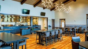 Un restaurant spacieux avec de hauts plafonds et des poutres en bois, comprenant un bar, plusieurs types de sièges et un éclairage moderne à suspension.
