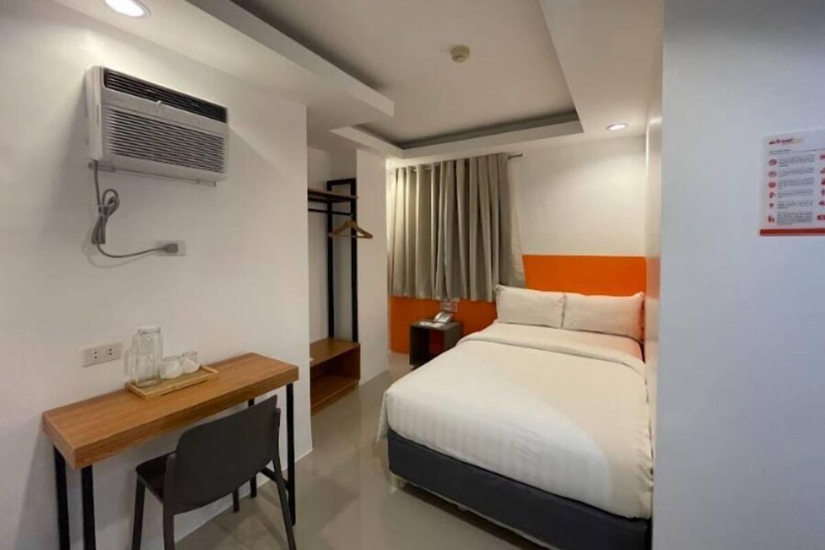 A hotel room with a bed, a desk, a chair, an air conditioning unit, and a coat rack.