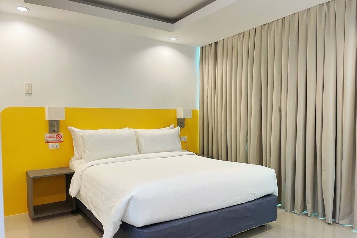 A modern hotel room with a bed, a nightstand, a wall-mounted light, and a curtain.