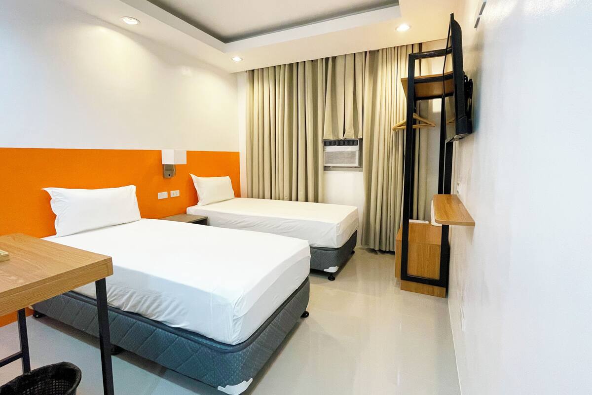 A hotel room with two beds, a desk, a TV mounted on the wall, and a window with curtains.