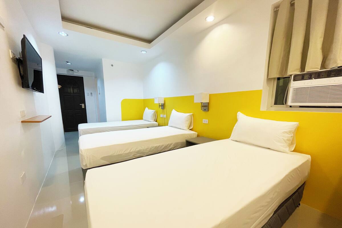A hotel room with three single beds, a flat-screen TV mounted on the wall, and a window with an air conditioning unit.