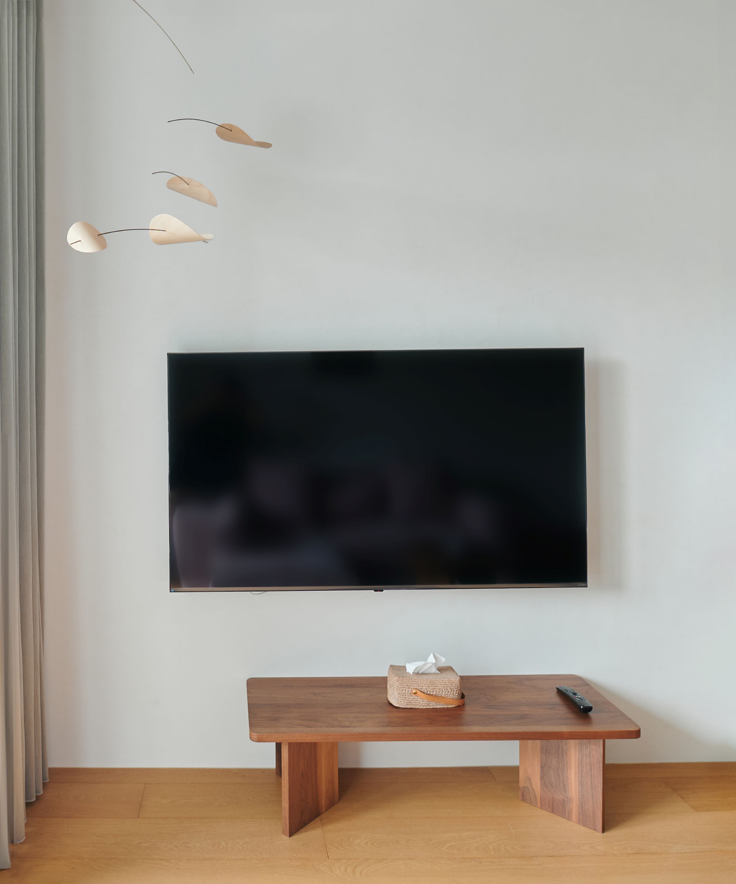 Un soggiorno moderno con TV a schermo piatto montata a parete, una panca di legno con scatola di fazzoletti e telecomando, e una lampada a sospensione semplice.