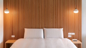 Un dormitorio moderno con cabecera de madera con listones, una cama grande con ropa de cama blanca, dos lámparas de noche y una mesita de noche con una lámpara y un enchufe.