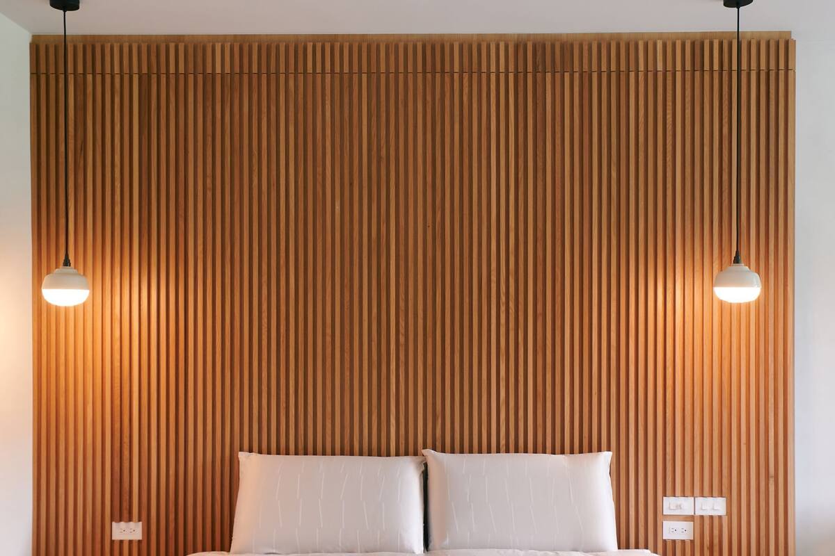 A modern bedroom with a wooden slatted headboard, a large bed with white linens, two bedside lamps, and a bedside table with a lamp and a power outlet.