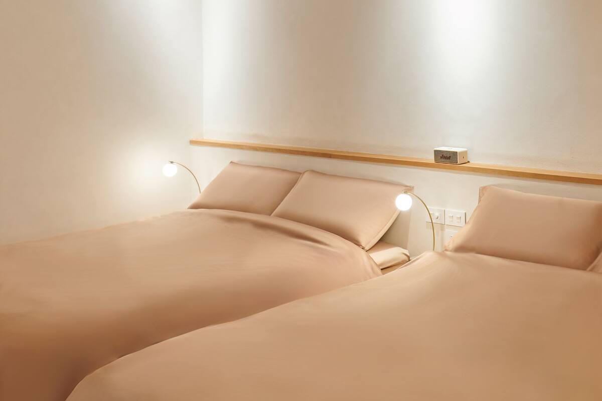 A minimalist hotel room with two beds, a bedside table, a lamp, and a shelf with a radio.