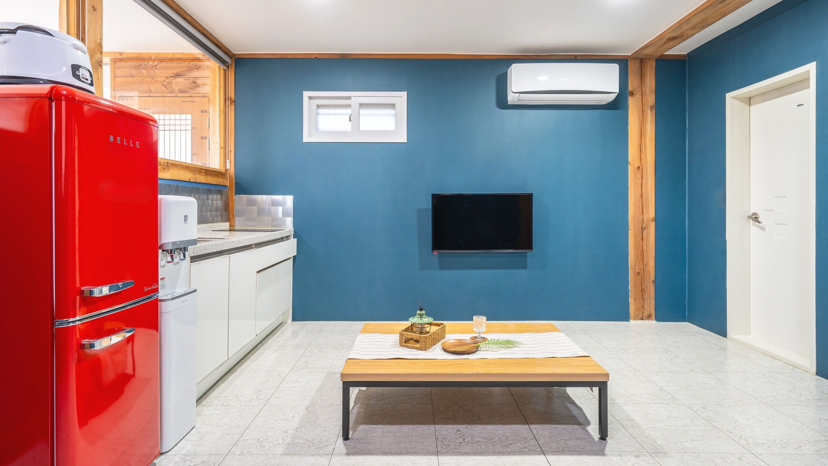 Cocina compacta con un refrigerador rojo, un microondas y un lavabo. Hay una mesita con una botella y un vaso. Las paredes son azules, y hay una puerta blanca y un equipo de aire acondicionado en la pared.