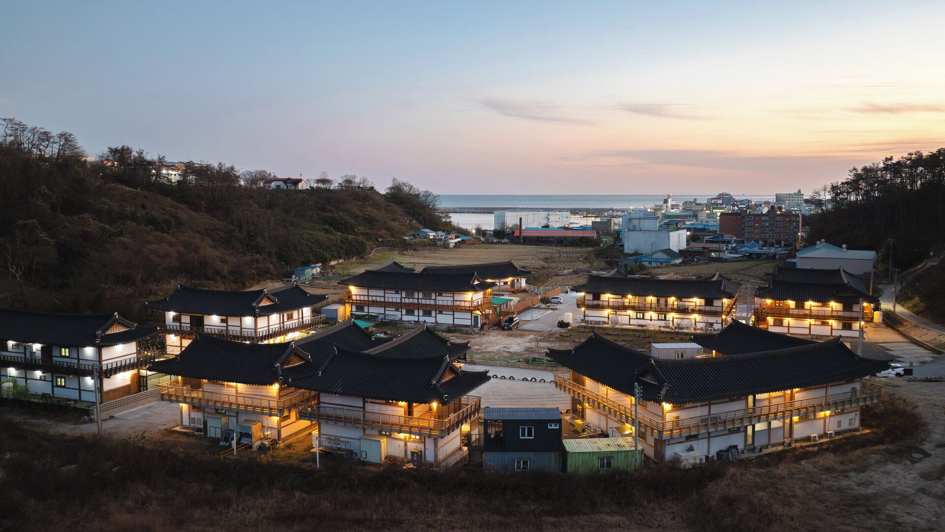 A traditional Japanese ryokan with multiple buildings, a dirt path, and a view of the sea and cityscape in the background.