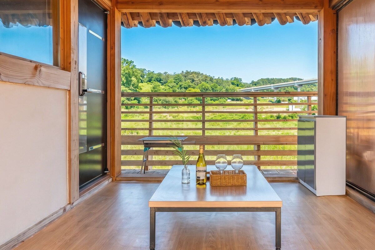 Une pièce avec une table en bois, une bouteille de vin et deux verres sur un balcon donnant sur un paysage verdoyant et un pont.
