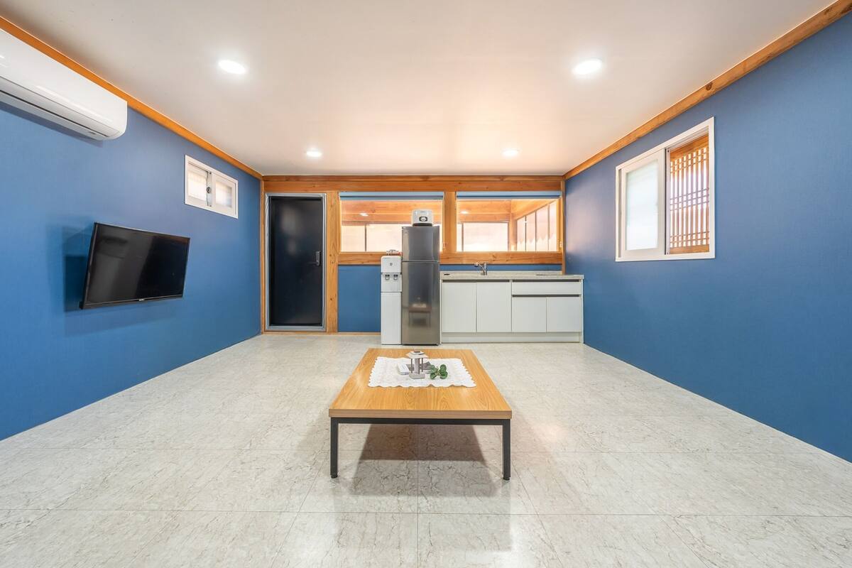 Ein Zimmer mit blauen Wänden, einer Holzbalkendecke, einem an der Wand befestigten Flachbildfernseher, einem kleinen Tisch mit einem dekorativen Gegenstand und einer Küchenzeile mit Kühlschrank und Spüle.