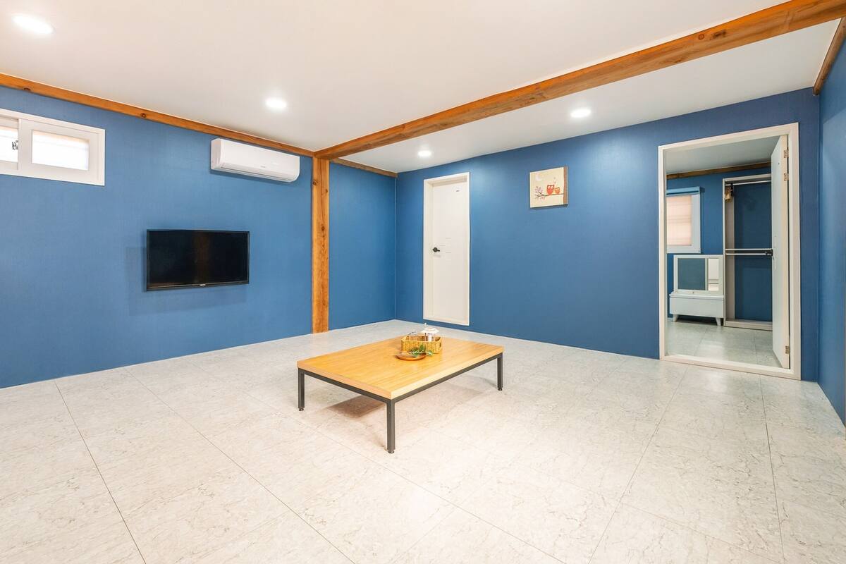 Ein Zimmer mit blauen Wänden, einer Holzbalkendecke, einem an der Wand montierten Flachbildfernseher, einem kleinen Holztisch mit Korb und Teller und einer Tür zu einem anderen Raum.
