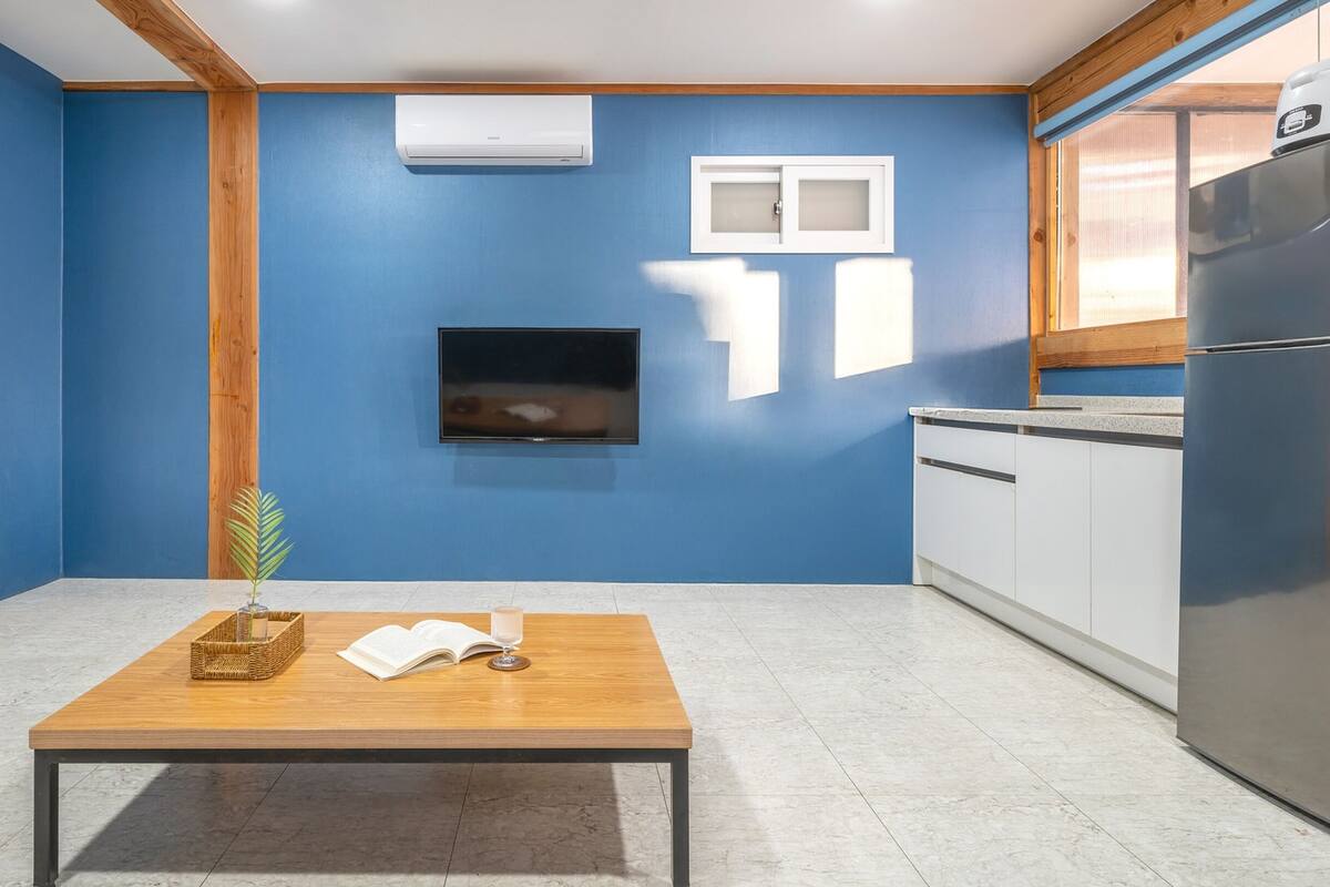 Eine kleine Küche mit blauen Wänden, einem Holztisch, einem Fernseher und einem Kühlschrank.