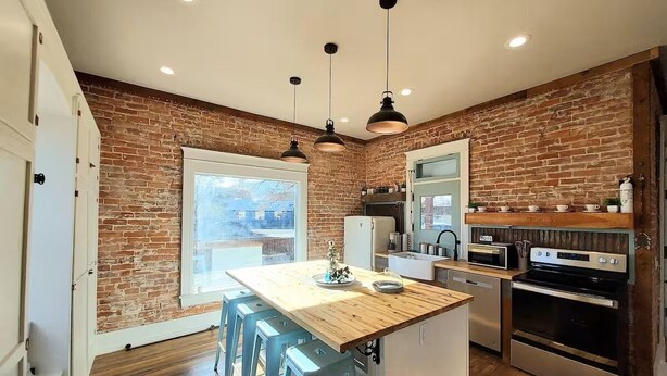 Una cocina con pared de ladrillo, taburetes de madera, una isla central y electrodomésticos modernos.