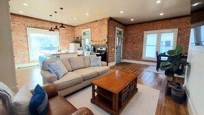 Amplia sala de estar con pared de ladrillo, mesa de centro de madera, sofá color beige y área de comedor con una planta grande.