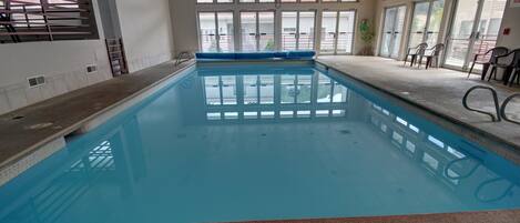 An indoor swimming pool with clear glass walls, a blue pool, and a seating area with chairs.