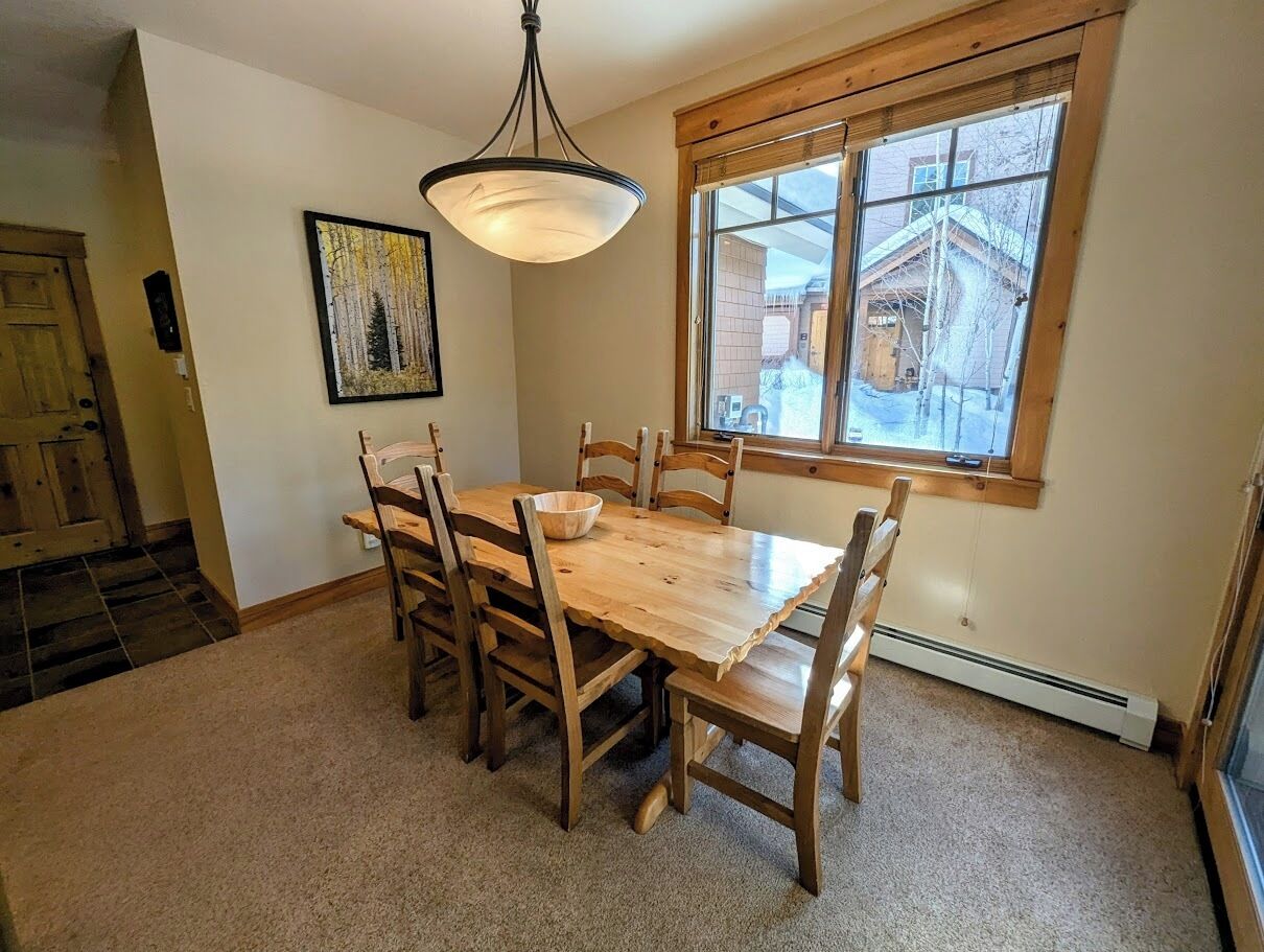 Un espace repas avec une table et des chaises en bois, un luminaire suspendu, une fenêtre donnant sur un bâtiment et une porte avec un cadre en bois.