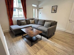 A living room with a sectional sofa, a coffee table, wooden flooring, a radiator, a door, and a wall-mounted picture.