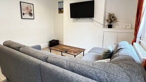 A living room with a grey sofa, a wooden coffee table, a flat-screen TV mounted on the wall, and a small potted plant.