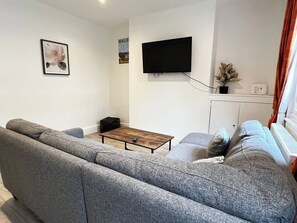 A living room with a grey sofa, a wooden coffee table, a flat-screen TV mounted on the wall, and a small potted plant.