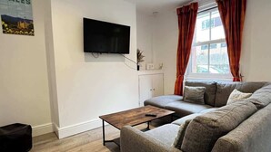 A living room with a sofa, a coffee table, a TV mounted on the wall, and a window with curtains.