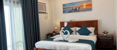 Un cuarto de hotel con una cama de madera, un cuadro de un atardecer, una ventana con cortinas y una mesita de noche con una lámpara.
