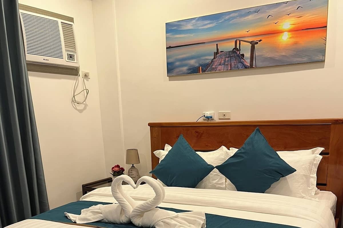 A neatly made bed with a wooden headboard, a painting of a sunset over water on the wall, and a pair of slippers on the floor.