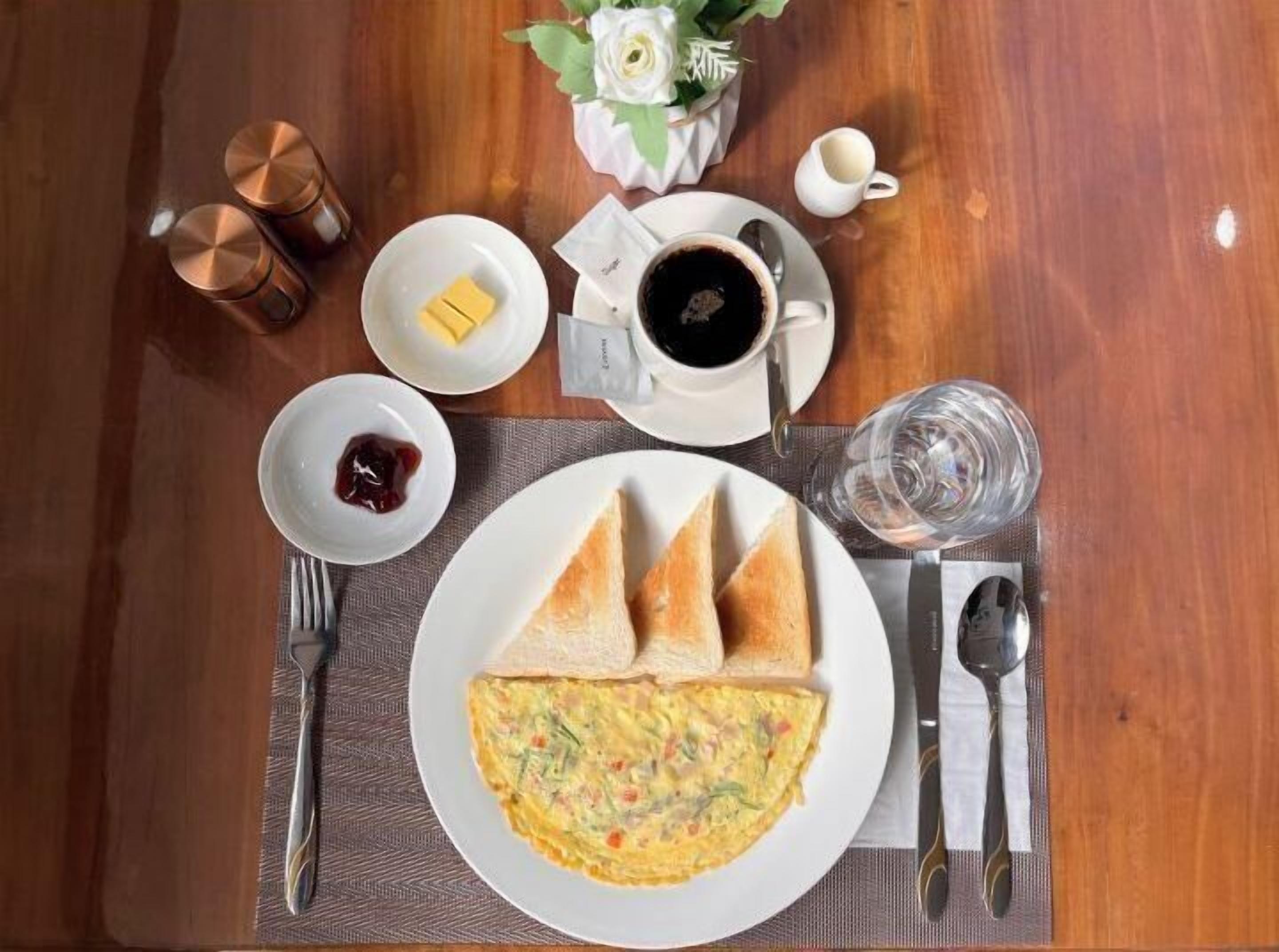 Un desayuno con tortilla, tostadas, mantequilla, mermelada, café y un vaso de agua sobre una mesa de madera.