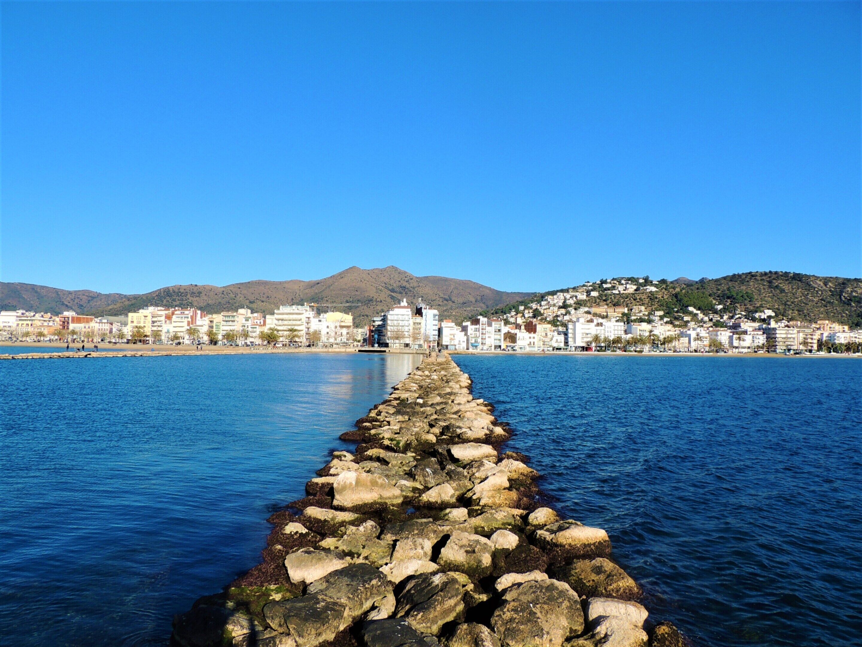A coastal cityscape with a prominent stone pier extending into the sea, a clear blue sky, and a backdrop of mountains and buildings.