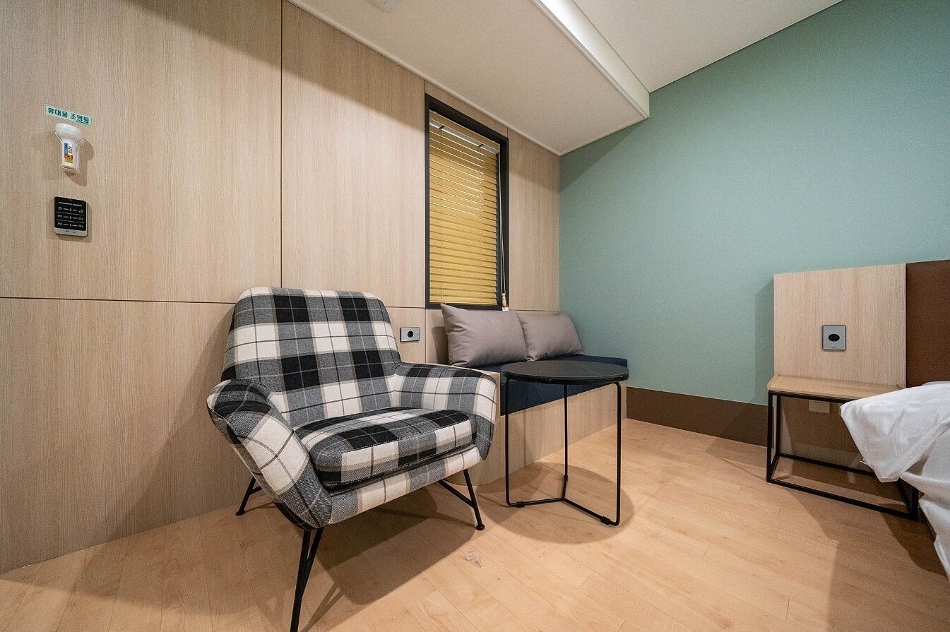 A compact hotel room with a plaid chair, a small table, a bed with a white cover, and a window with blinds.