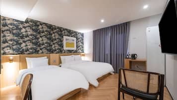 A hotel room with two beds, a wooden floor, a rattan chair, a TV, and a wall with floral wallpaper.