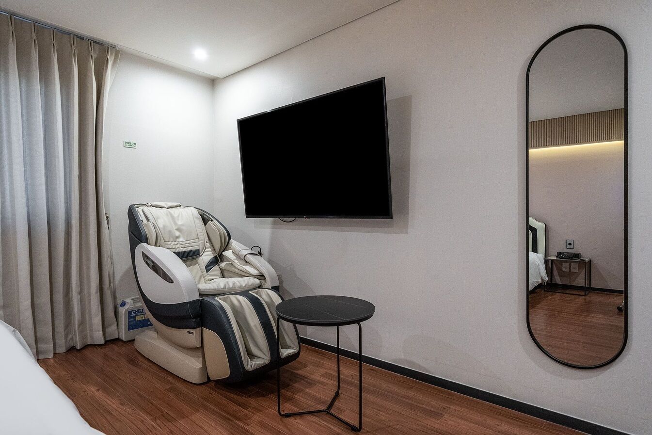 A room with a massage chair, a flat-screen TV, a small table, and a large mirror.