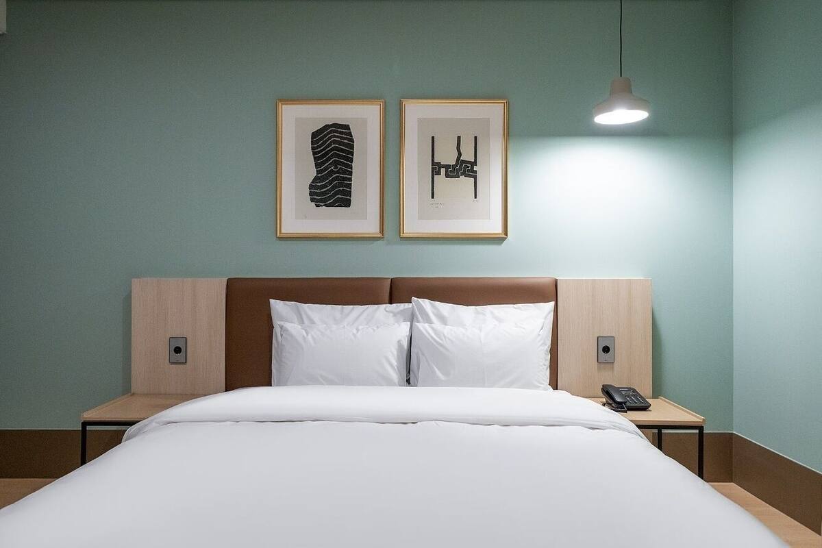 Une chambre d’hôtel avec un lit, deux œuvres d’art encadrées au mur, une table de chevet avec un téléphone et un luminaire suspendu.