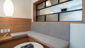Une chambre d’hôtel moderne avec une tête de lit en bois, un canapé gris, une table ronde avec un coffre intégré et une grande fenêtre avec des stores.