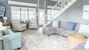 A spacious living room with a grey sectional sofa, striped armchairs, a white coffee table, and a patterned rug. A staircase with white railings leads to an upper level.