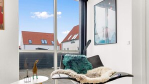 Ein modernes Wohnzimmer mit großem Fenster, einem schwarzen Sessel mit flauschigem Kissen, einem kleinen runden Tisch und einem gerahmten Bild an der Wand.