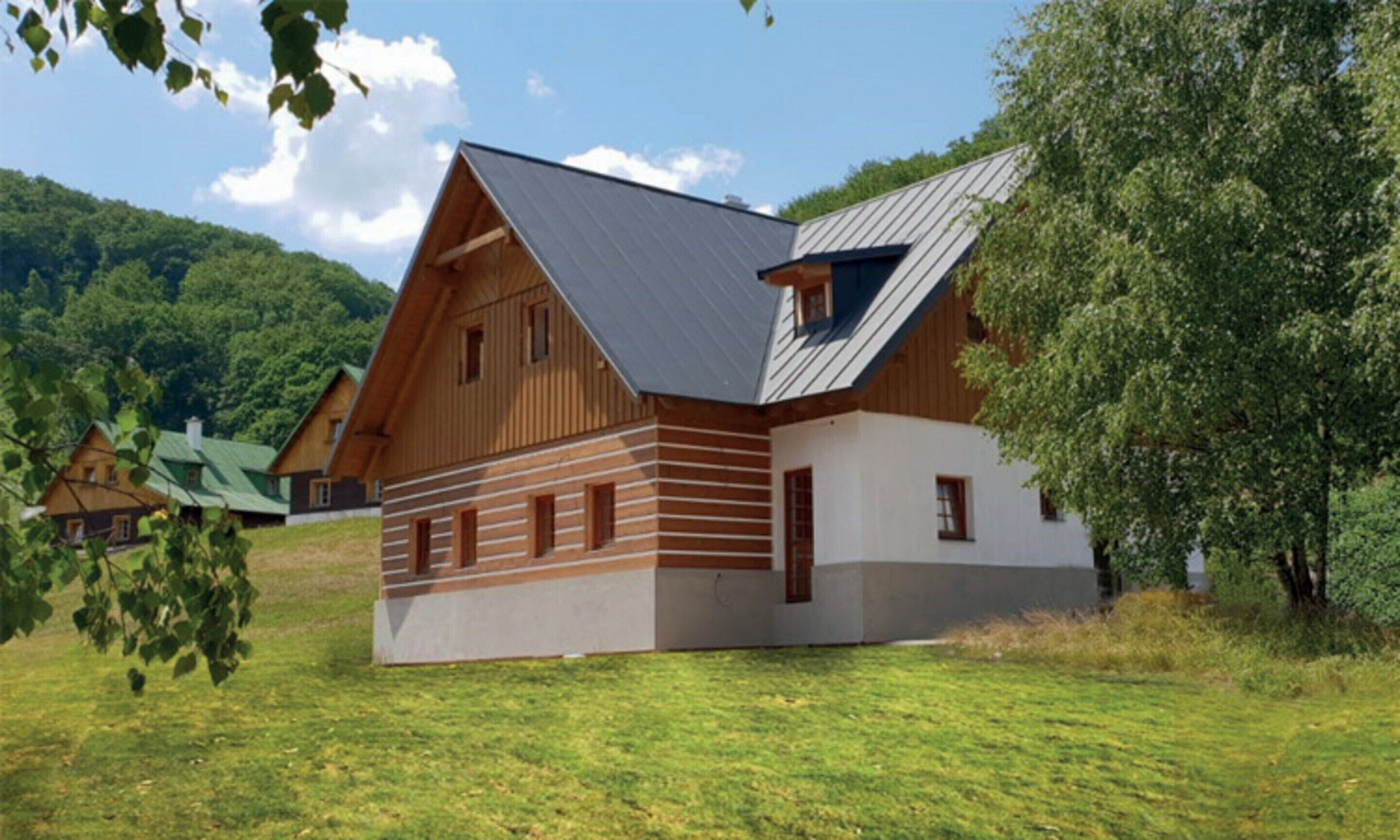 A modern wooden house with a sloped roof and large windows, surrounded by a grassy field and trees.