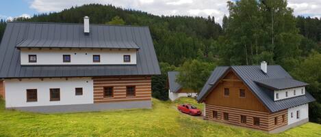 Una casa a due piani con un tetto spiovente, un'auto parcheggiata di fronte e un pendio boscoso sullo sfondo.