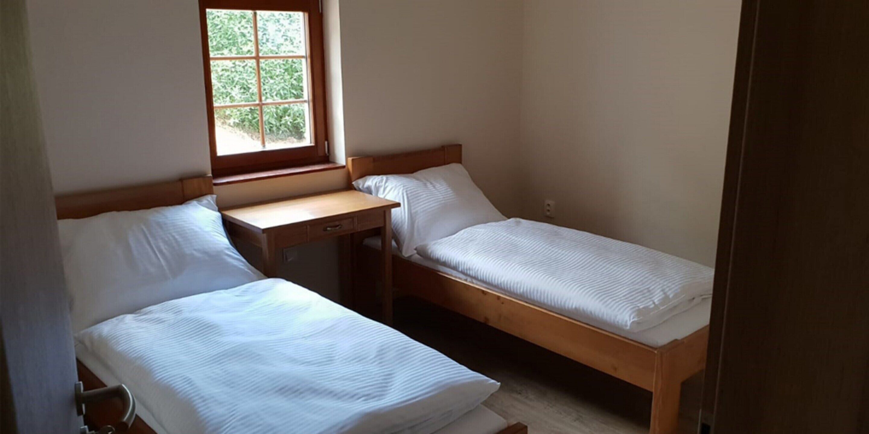 Eine kleine, einfache Unterkunft mit zwei Einzelbetten, einem Holztisch und einem Fenster mit Tageslicht.