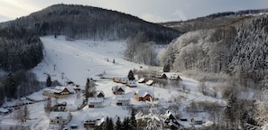 Uma aldeia coberta de neve com casas de madeira tradicionais, um teleférico de esqui e uma montanha arborizada ao fundo.