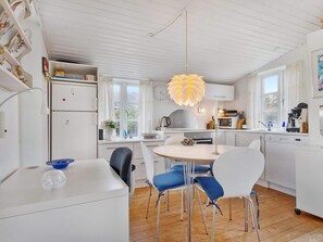 A modern kitchen with white cabinetry, a dining area with a round table and blue chairs, and a unique leaf-shaped pendant light. - 5 Star Holiday Home in Lokken (Lokken)