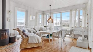 A cozy room with a fireplace, wicker furniture, a dining table, and large windows offering a view of the outdoors. - 5 Star Holiday Home in Lokken (Lokken)