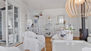 A white-washed interior with a large window, wicker furniture, a white table, and a wood-burning stove. - 5 Star Holiday Home in Lokken (Lokken)