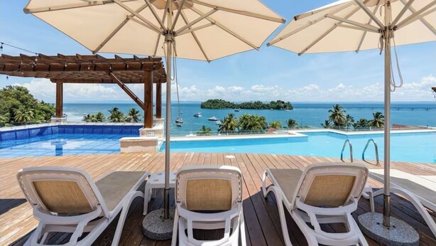 A poolside area with lounge chairs and umbrellas, overlooking a clear blue sea with boats and an island in the distance. - Rent a home away from home in front of the Best View of beautiful Samaná Bay! (Samana)