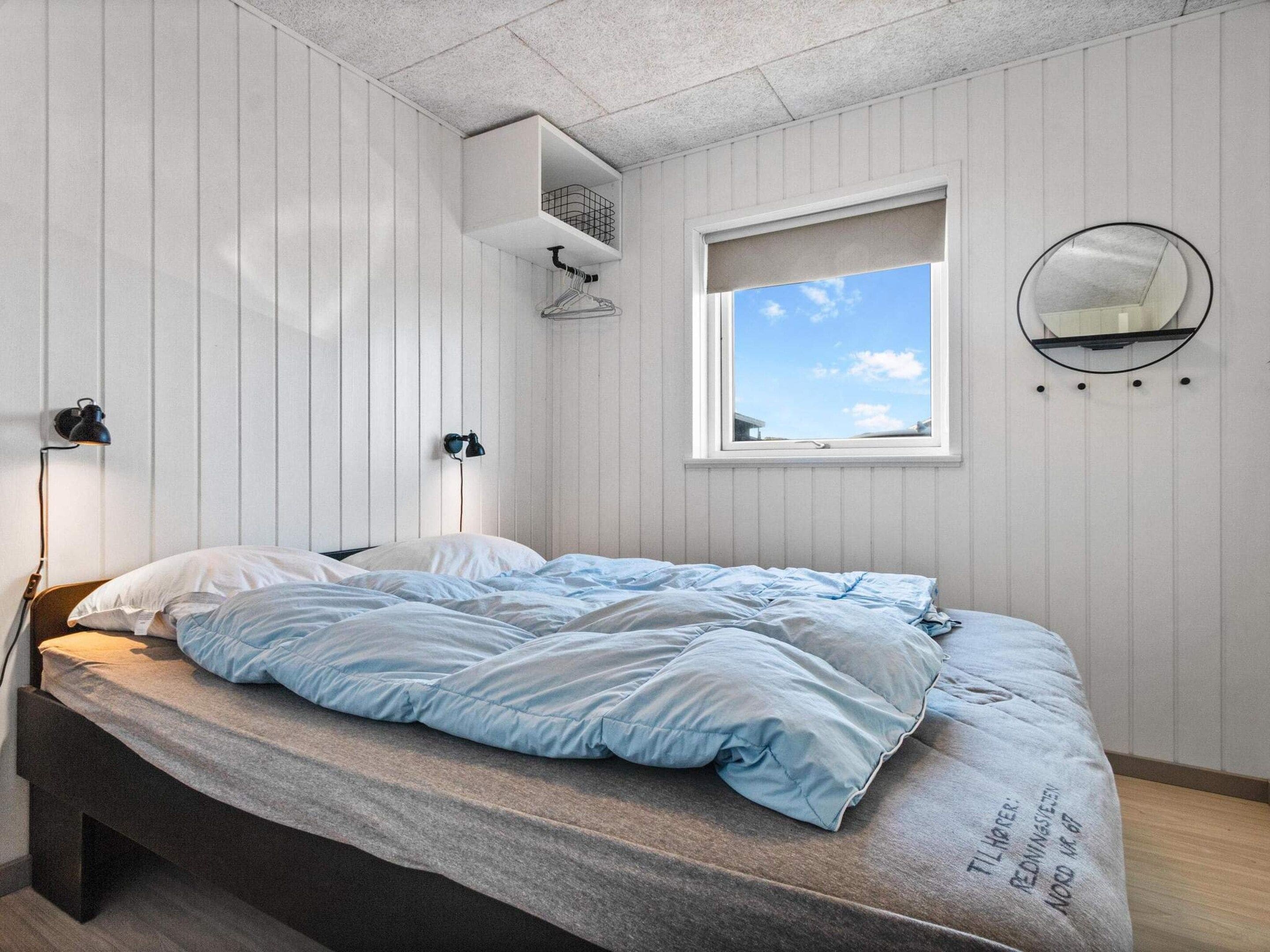 A bedroom with a bed, a window, a wall-mounted mirror, and a shelf with a basket.