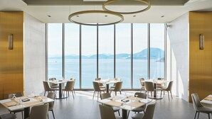 A modern dining area with tables set for four, large windows offering a view of the sea and distant mountains, and a ceiling with circular light fixtures. - YEOSU BELLE MER (Yeosu)