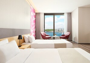A hotel room with a large bed, two red chairs, a small table, and a view of the city and water.