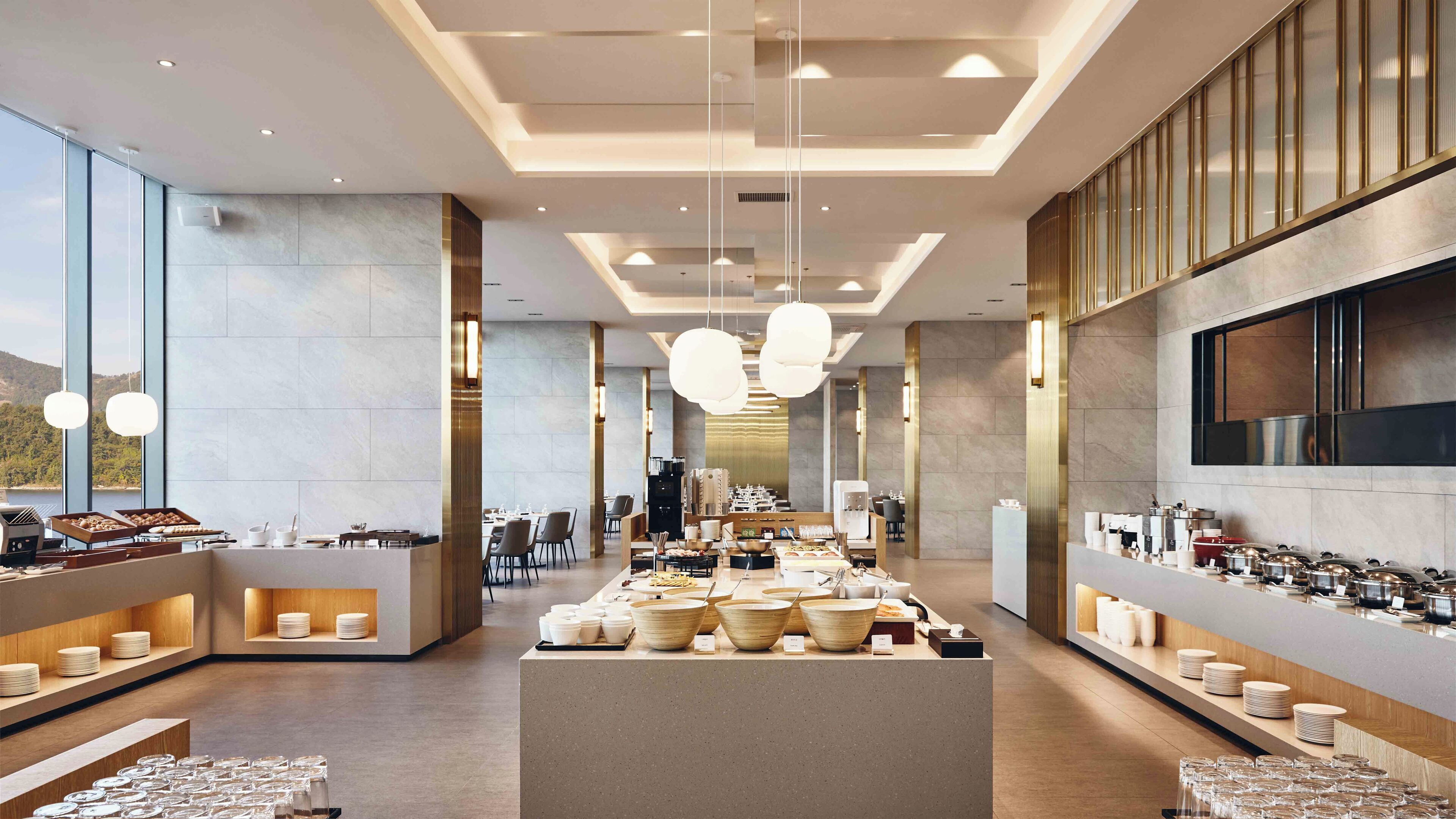 A modern hotel dining area with a central buffet, long tables, and a view of the outdoors.